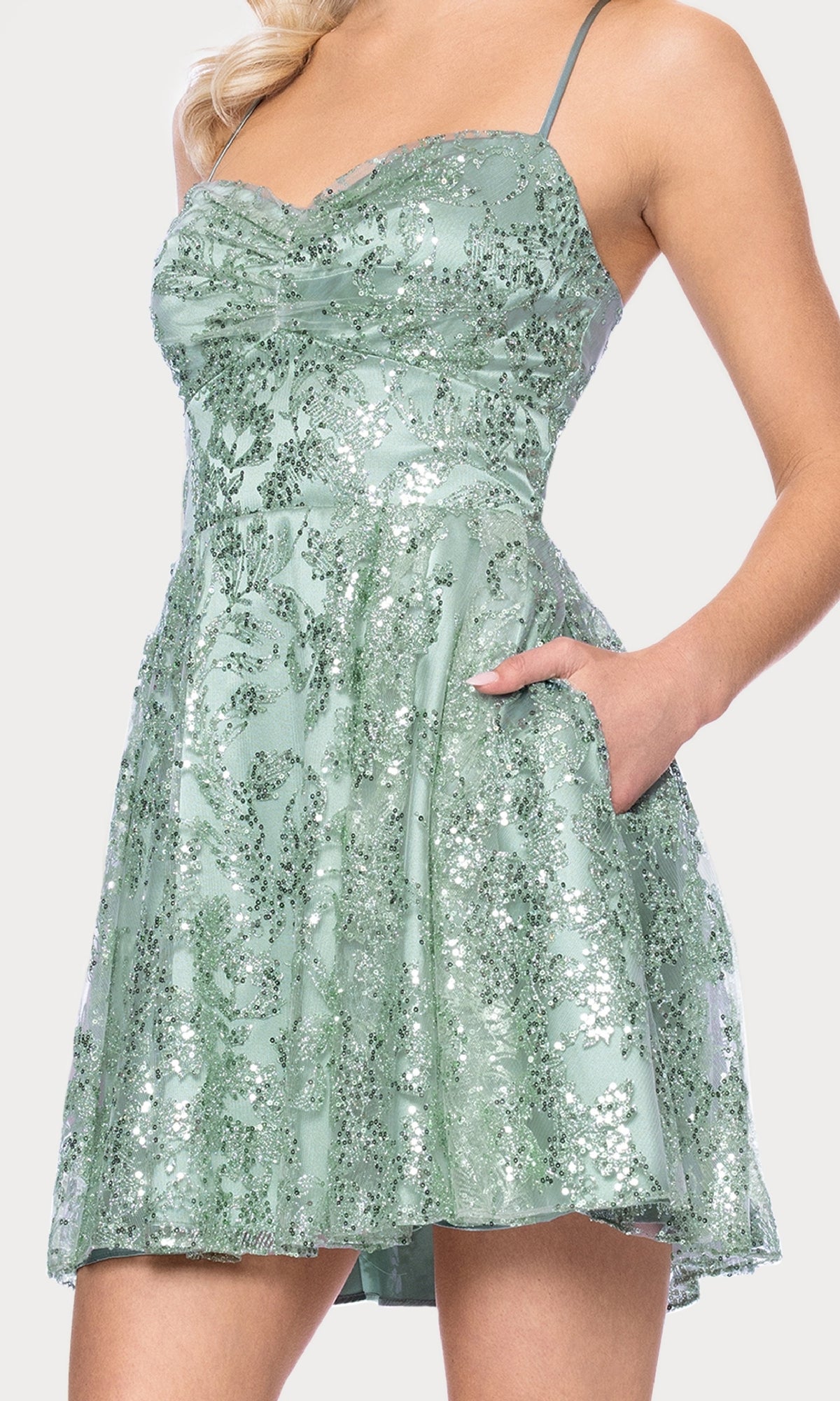 A woman confidently poses with one hand in the pocket of the Mini Green Sequin Hoco Dress: Blondie Nites 5705BN, featuring thin straps and a flared skirt—perfect for junior sizes.